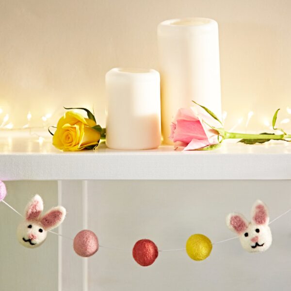 Candles and roses adorn a white shelf, while the delightful Felt Bunny Garland is draped below, featuring a charming felt bunny surrounded by colorful pom-poms.
