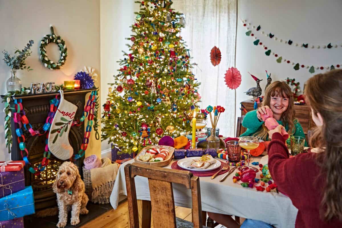 Two women sit at a decorated dining table in a festively adorned room, with Christmas decorations, a Christmas tree, stockings on the fireplace, and a festive Christmas banner whilst their dog rests nearby.