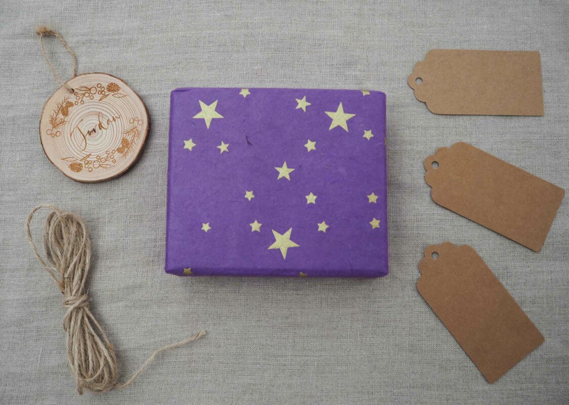 A purple gift wrapped with yellow stars is displayed on a gray fabric background, accompanied by twine, a round wooden ornament made from elephant dung, and three blank brown gift tags.