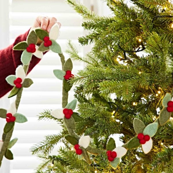 A person in a red sleeve decorates a Christmas tree with the Felt Holly Christmas Garland, featuring felt leaves and red berries.