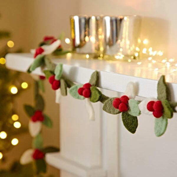 A fireplace adorned with the Felt Holly Christmas Garland (Copy), accompanied by silver candle holders and string lights, with a lit Christmas tree in the background.