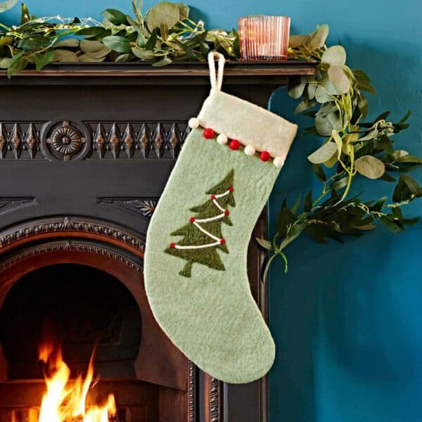 A green felt Christmas stocking with a tree design hangs on a black fireplace mantel decorated with greenery, whilst a lit candle glows and a fire burns warmly in the hearth below.
