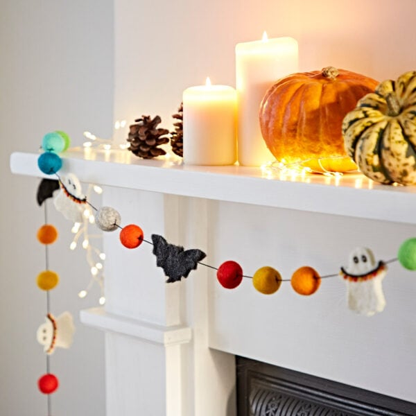 A white mantel decorated with pumpkins, pinecones, candles, and vibrant Halloween decor featuring the Felt Ghosts and Bats Garland, with string lights illuminating the scene.