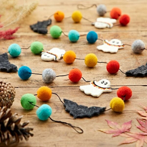The Felt Ghosts and Bats Garland, displaying vibrant pom-poms, felt ghost and bat shapes, is elegantly showcased on a wooden surface accented with autumn leaves and pine cones in the background.