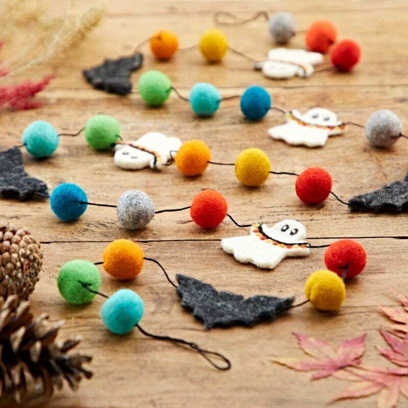 The Felt Ghosts and Bats Garland, displaying vibrant pom-poms, felt ghost and bat shapes, is elegantly showcased on a wooden surface accented with autumn leaves and pine cones in the background.