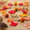A Felt Pumpkin and Leaves Garland, showcasing a charming felt pumpkin with jack-o'-lantern faces and an array of vibrantly colored leaves garland, is laid on a wooden surface. Surrounding the garland are scattered pinecones and dried plants.