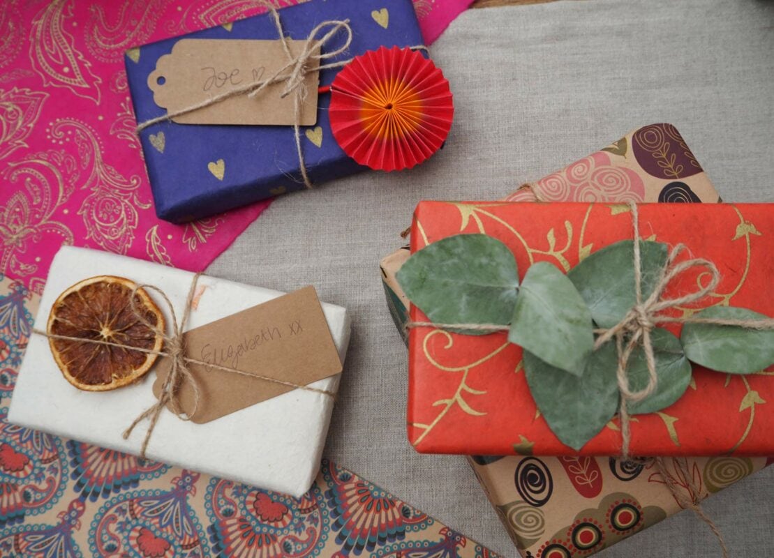 Three wrapped gifts with labels are arranged on a colorful, patterned cloth. The gifts are decorated with twine, a dried citrus slice, a red paper rosette, leaves, and an eco-friendly touch using elephant dung paper.