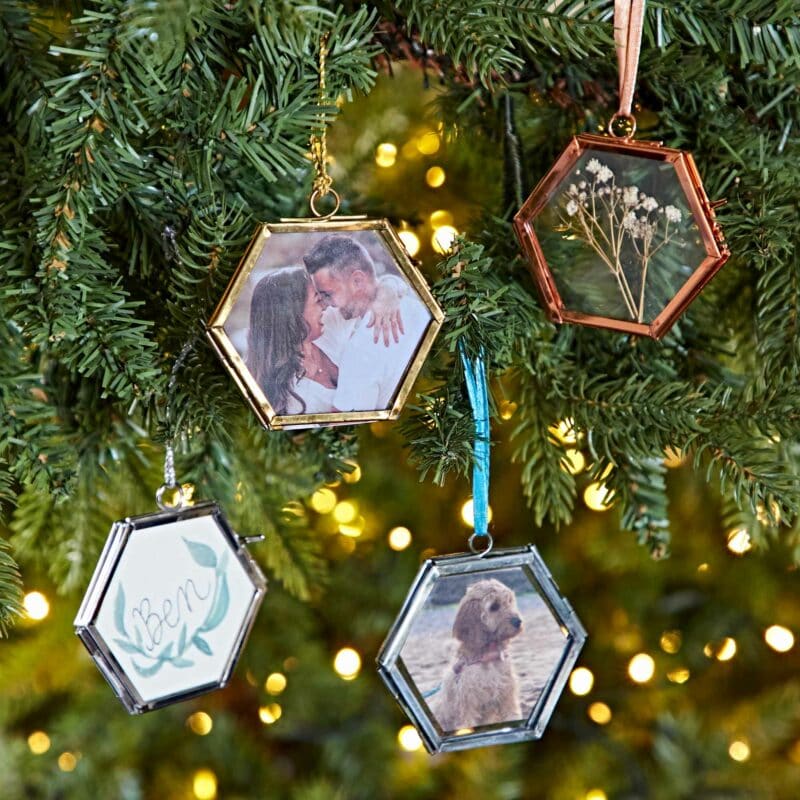 Close-up of a decorated Christmas tree branch featuring four hexagonal ornaments, each containing a unique insert: a couple's photo, dried flowers, a written name, and a dog photo. These charming decorations make perfect Autumn Style Gifts.