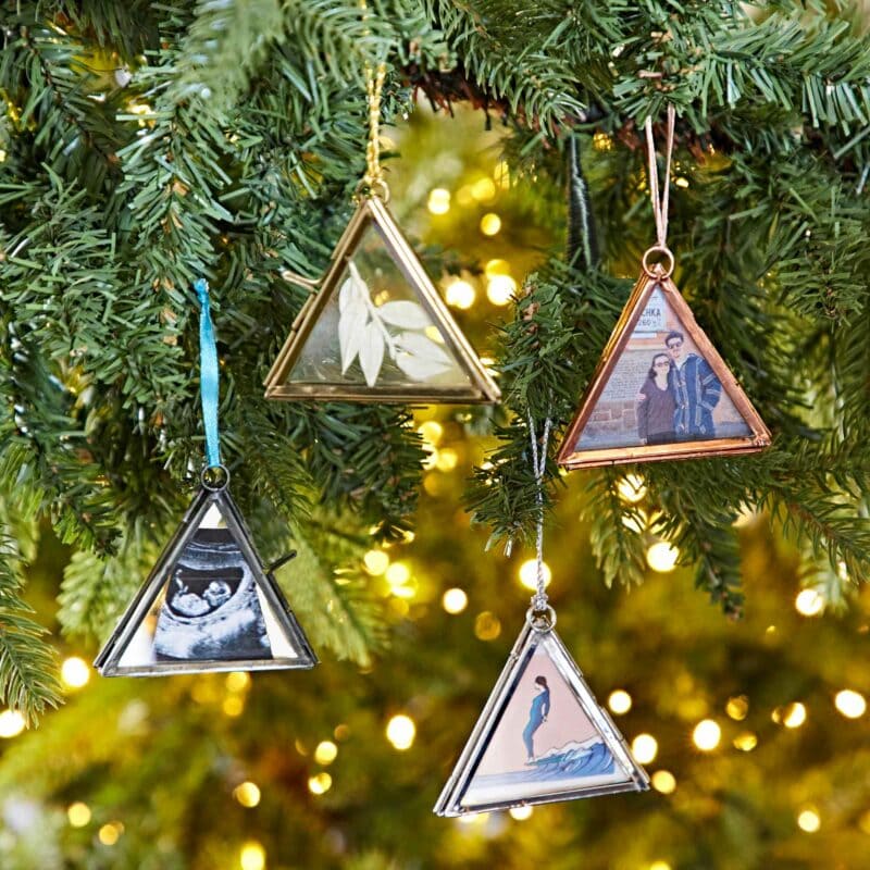 Four triangular photo ornaments, perfect as Autumn Style Gifts, hang on a Christmas tree. Each ornament contains a different image and is framed in various metallic colors. The background shows green branches and twinkling lights.