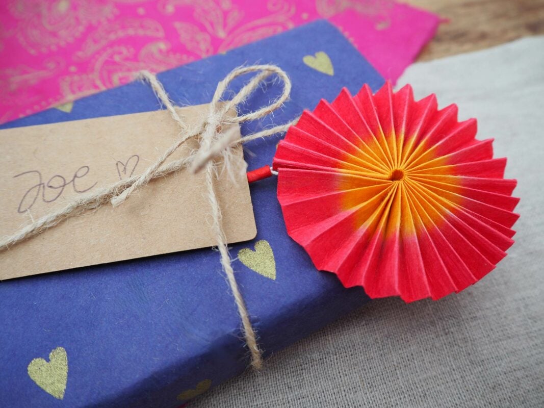 A wrapped gift with blue paper decorated with gold hearts, a brown tag labeled "Zoe" with a heart, and a red and yellow decorative pinwheel attached. It sits on an exotic cloth surface made from elephant dung fibers.