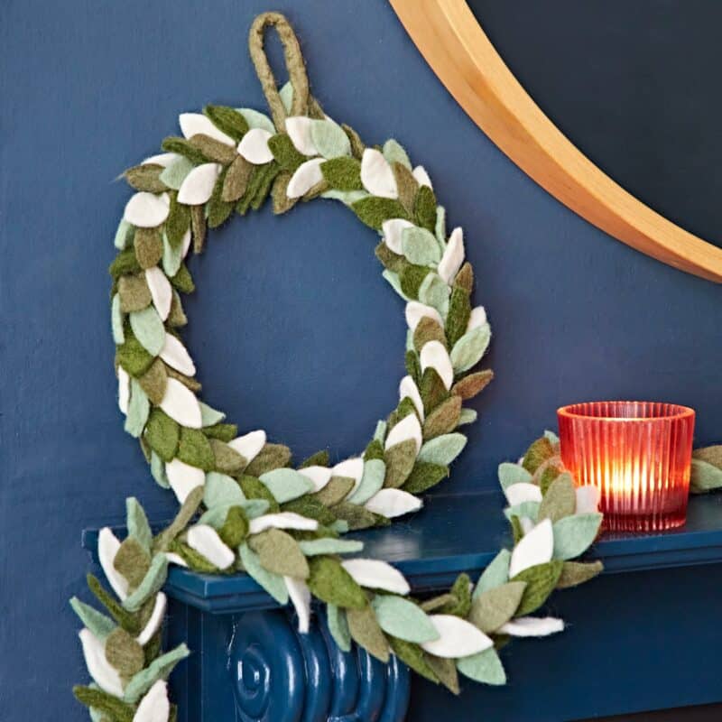 A felt leaf wreath and Christmas bunting garland in green and white are displayed on a dark blue mantelpiece next to a lit red glass candle holder, creating a festive Christmas decoration.