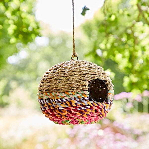 The Large Round Recycled Cotton Birdhouse is woven with recycled cotton fibers, boasts a natural design with multicolored accents, and is shown hanging outdoors against a blurred green background.