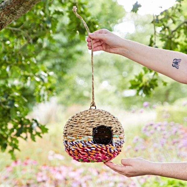 A person hangs a Large Round Recycled Cotton Birdhouse, featuring colorful stripes, from a tree branch.