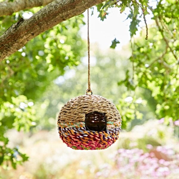 The Large Round Recycled Cotton Birdhouse, adorned with a colorful band, hangs gracefully from a tree branch among lush greenery.