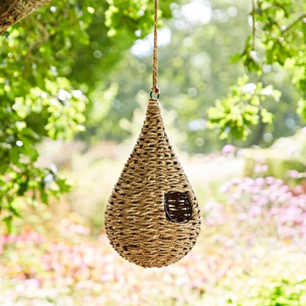 The Teardrop Jute Birdhouse, crafted from jute, hangs elegantly from a branch, adding rustic allure to the garden.