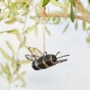 The Recycled Metal Hanging Bee Garden Ornament features detailed wings and hangs from a tree branch by a string, set among green leaves with a softly blurred background.
