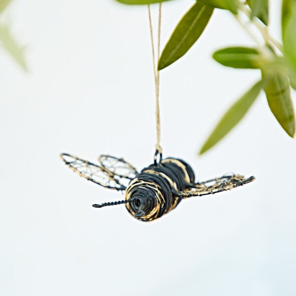 The Recycled Metal Hanging Bee Garden Ornament features a black and gold bee crafted from recycled metal, hanging by a string on a branch with green leaves in the background.
