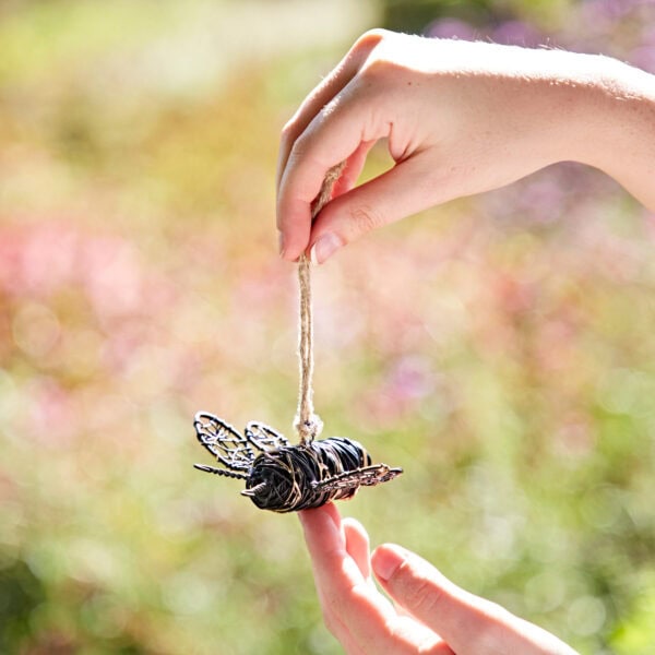 A hand holds a Hanging Garden Yellow and Black Bee Ornament against a blurred outdoor background.