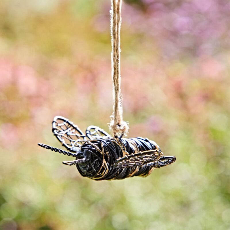 A yellow and black bee ornament, crafted from twisted wire, hangs like a delicate treasure in a garden, suspended on a string against a blurred natural background.