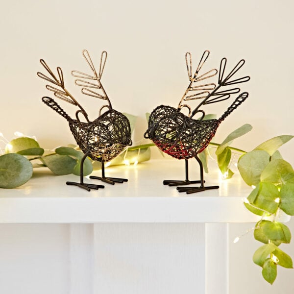 The Set of Two Small Wire Robins with Wings Decorative Ornament features bird sculptures with leaf-like antlers and delicate wings on a white surface, surrounded by eucalyptus leaves and string lights, adding whimsical charm to any space.