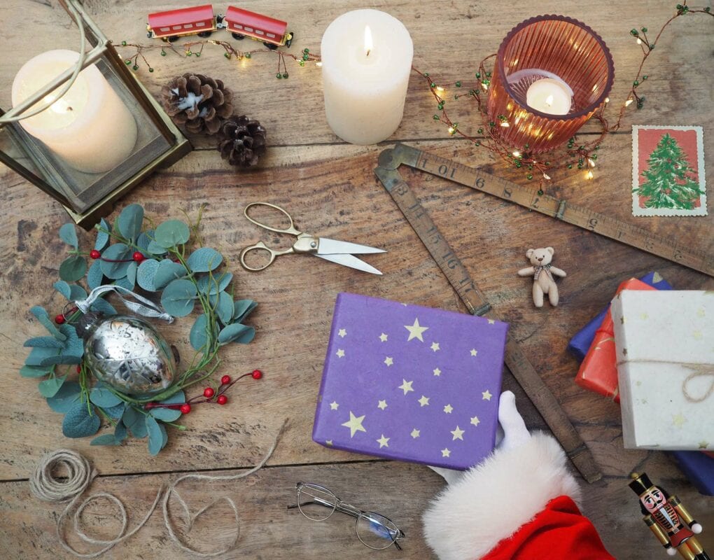 A festive tabletop showcases a wrapped gift, Santa's hand, candles, scissors, a decorative sprig, a wooden ruler, a toy train crafted from recycled materials like elephant dung paper, and twinkling holiday lights.