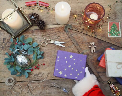 A festive tabletop showcases a wrapped gift, Santa's hand, candles, scissors, a decorative sprig, a wooden ruler, a toy train crafted from recycled materials like elephant dung paper, and twinkling holiday lights.