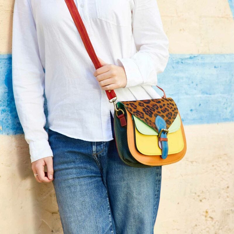 A person in jeans and a white shirt holds a colorful saddle crossbody bag featuring a leopard print flap and bold red strap.