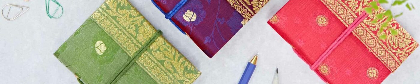 Three cloth-bound notebooks in green, purple, and red with ornate designs are arranged on a surface with a pen and paperclips nearby—an unusual gift for those who cherish the art of writing.