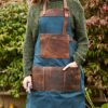 A person stands outdoors, sporting a green canvas sweater complemented by the Personalised Green Canvas and Buffalo Leather Apron, which features blue fabric with brown leather pockets. The apron, crafted from high-quality buffalo leather, adds a personalized touch to their ensemble.