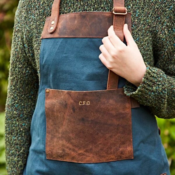 A person is adjusting the strap of a Personalised Green Canvas and Buffalo Leather Apron, featuring brown buffalo leather pockets and straps, embroidered with the initials "C.F.G." over a green sweater.