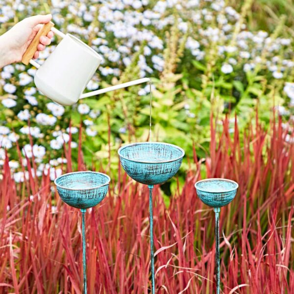 A white watering can pours water over three metallic turquoise garden ornaments, featuring a Pantala Metal Dragonfly Garden Stake Decoration, amid red and green foliage.