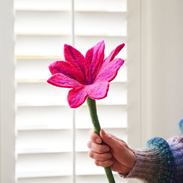 A hand holding a vibrant fuchsia felt flower in front of white shutters.