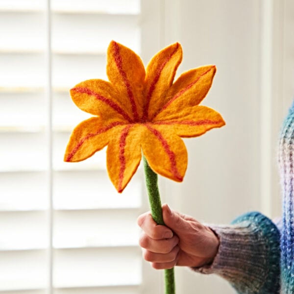 A person holds a handmade felt flower with mustard-orange petals and red veins against a backdrop of white window shutters.