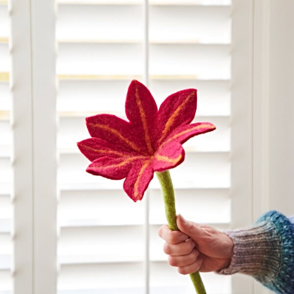 A hand holds a red felt flower with a green stem, its vibrant hue reminiscent of a rich wine, set against the backdrop of white window shutters.