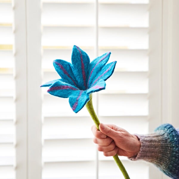 A person holding a turquoise felt flower in front of white window shutters.
