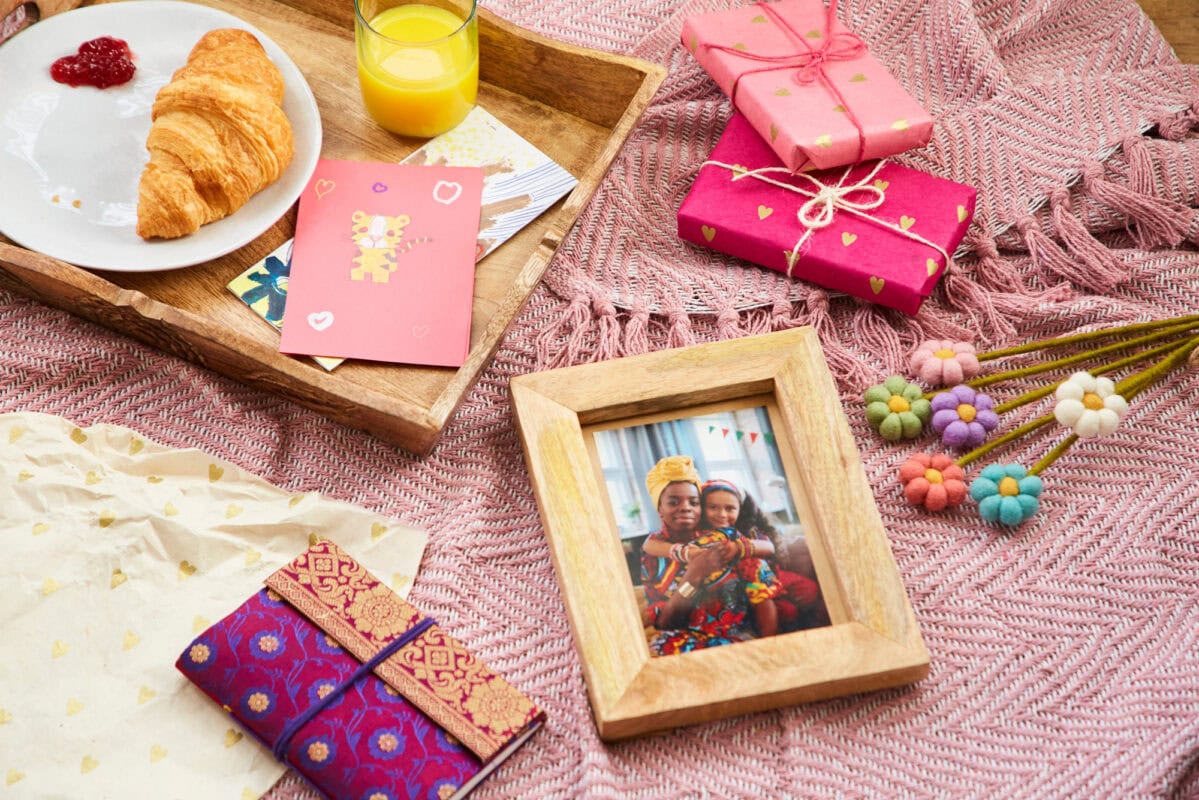 A tray with a croissant, jam, orange juice, and a Mother's Day greeting card sits on a fabric. Nearby are wrapped gifts, a photo frame, colorful flowers, and decorative envelopes—creating the perfect Mother's Day gift setting.