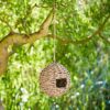 The Round Munja Birdhouse, made from woven straw, hangs from a tree branch with green foliage in the background.