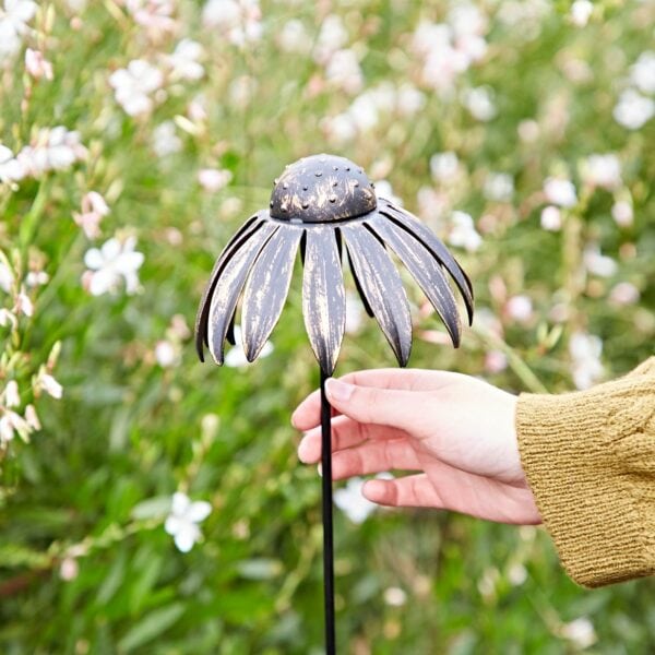 A hand holds a Metal Echinacea Flower Garden Stake Decoration in a garden with blooming white flowers in the background.