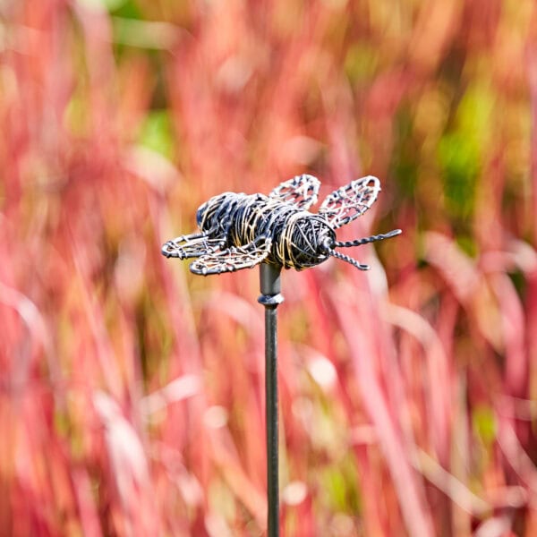 A hanging yellow and black bee garden ornament on a thin pole stands against a backdrop of tall, blurred reddish-pink grass.