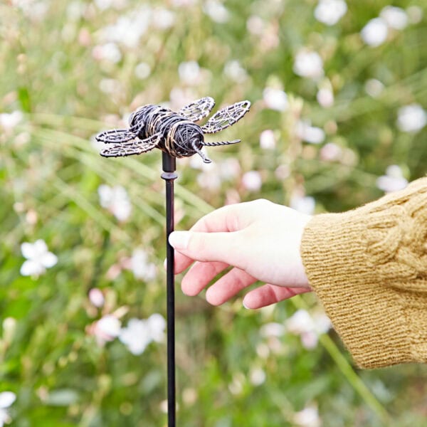 A hand reaches for a Hanging Yellow and Black Bee Garden Ornament on a stick, set against green plants and white flowers.
