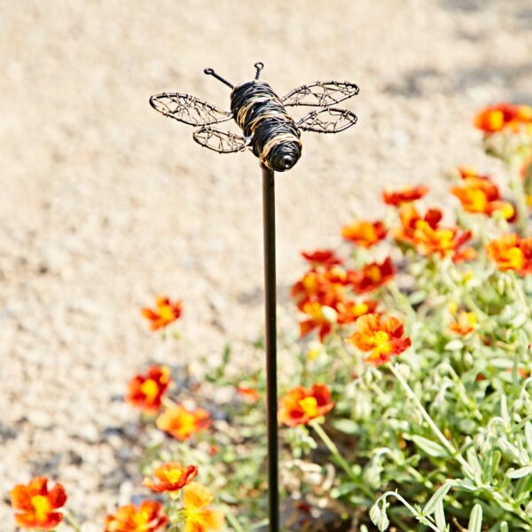 The Recycled Metal Bee Garden Stake features a bee on a slim black pole, creating unique garden decor among orange and yellow flowers with a gravel backdrop.