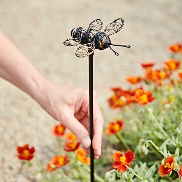 Someone places the Recycled Metal Bee Garden Stake into the soil beside blooming orange and yellow flowers.