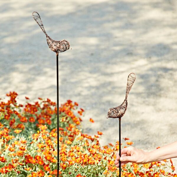 Two Recycled Metal Bird Garden Stake Decorations are placed among orange flowers; a hand adjusts the shorter stake.