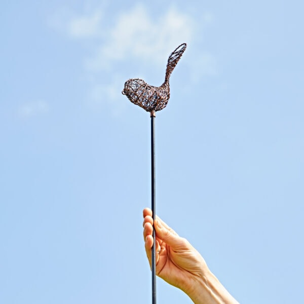A hand holds up the Recycled Metal Bird Garden Stake Decoration, featuring a bird crafted from recycled metal on a thin rod, set against a clear blue sky.