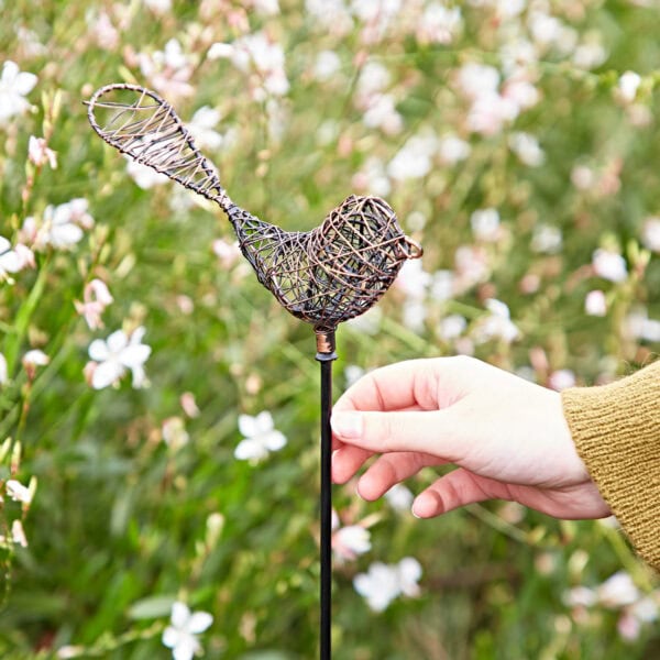 In a garden adorned with blooming plants, a hand grasps a Metal Bee Garden Stake Ornament, exemplifying a delightful accent to enrich your garden's natural allure.