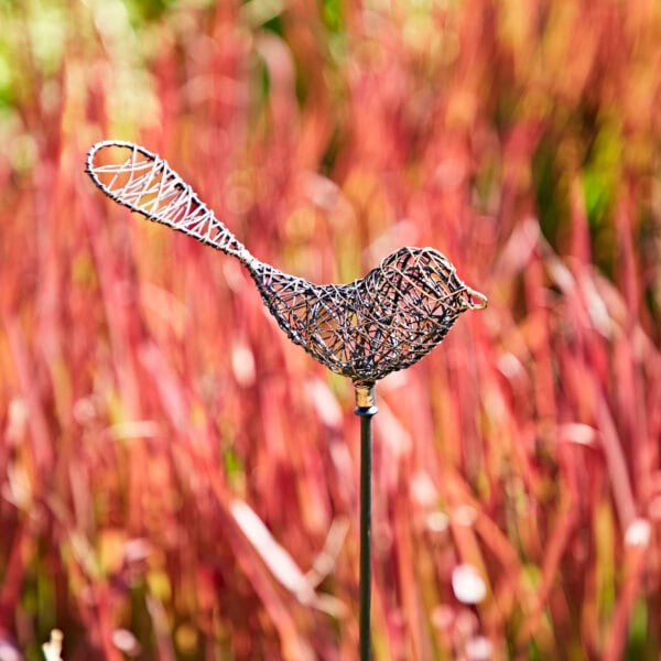 The Metal Bee Garden Stake Ornament pairs beautifully with a wire bird sculpture on a pole, set against vibrant red and green plants, creating a charming garden display.