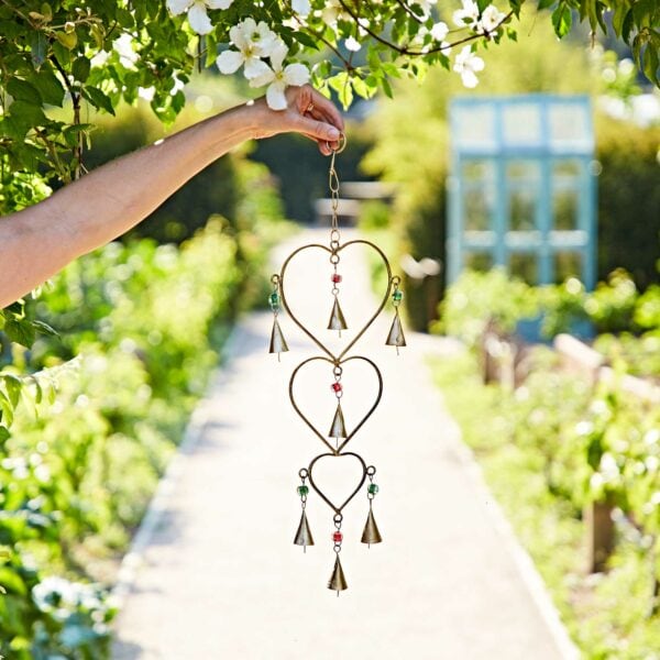 A hand holds the Recycled Iron Heart and Bells Wind Chime, featuring heart shapes and small bells, in a sunny garden with green plants and a pathway behind.