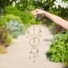 A person holds the Recycled Iron Heart and Bells Wind Chime, featuring heart and bell shapes with beads, outdoors in a garden setting.