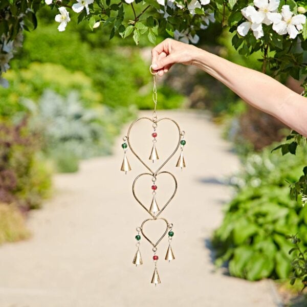 A person holds the Recycled Iron Heart and Bells Wind Chime, featuring heart and bell shapes with beads, outdoors in a garden setting.