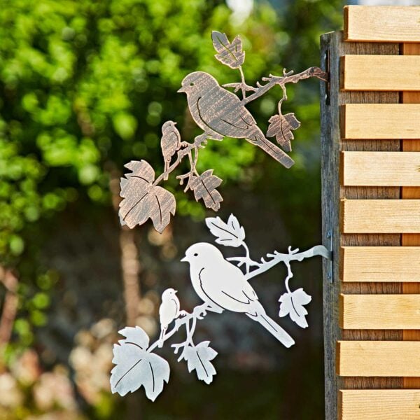 Two Recycled Iron Bird Silhouette Garden Art pieces, one silver and one brown, are displayed on a wooden fence with lush greenery in the background.